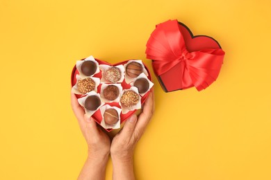 Photo of Woman with heart shaped box of delicious chocolate candies on yellow background, top view