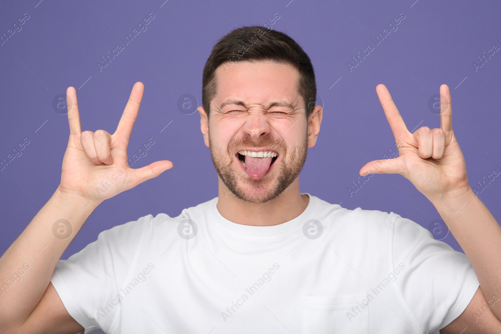 Photo of Happy man showing his tongue and rock gesture on purple background