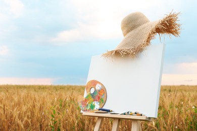 Photo of Wooden easel with blank canvas, painting equipment and hat in field. Space for text