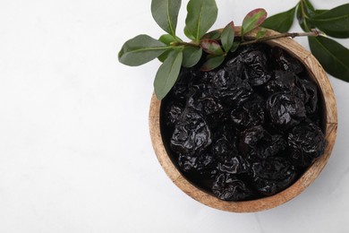 Sweet dried prunes in bowl and green leaves on white table, top view. Space for text