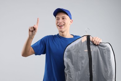 Dry-cleaning delivery. Happy courier holding garment cover with clothes and pointing at something on light grey background