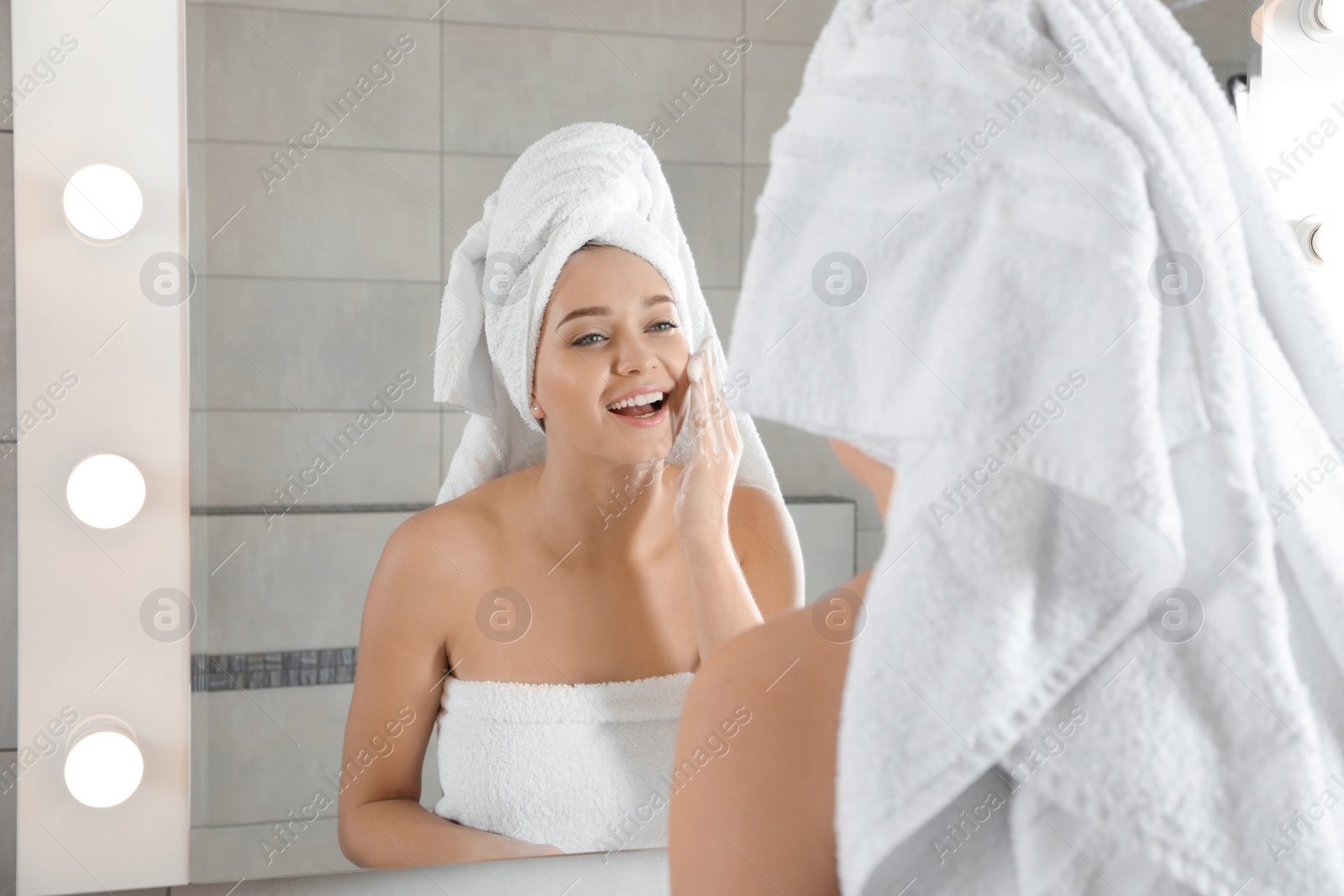 Photo of Young woman washing face with soap near mirror in bathroom