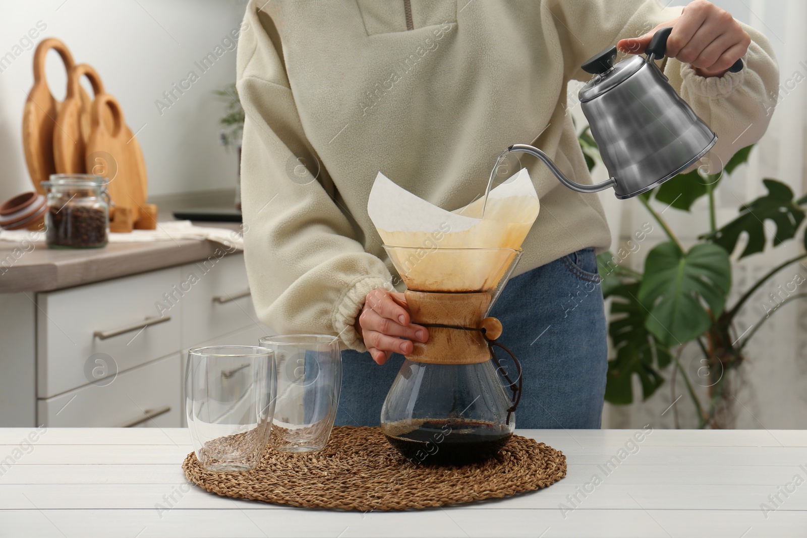 Photo of Woman pouring hot water into glass chemex coffeemaker with paper filter and coffee at white table, closeup