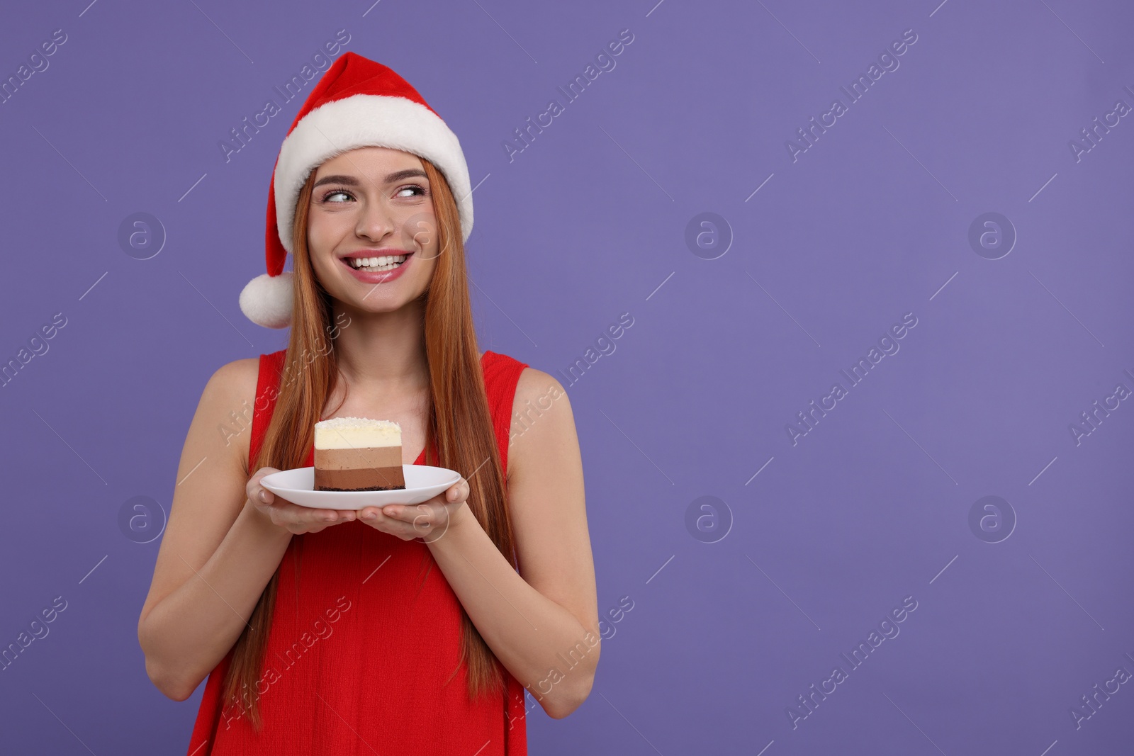 Photo of Young woman in Santa hat with piece of cake on purple background, space for text