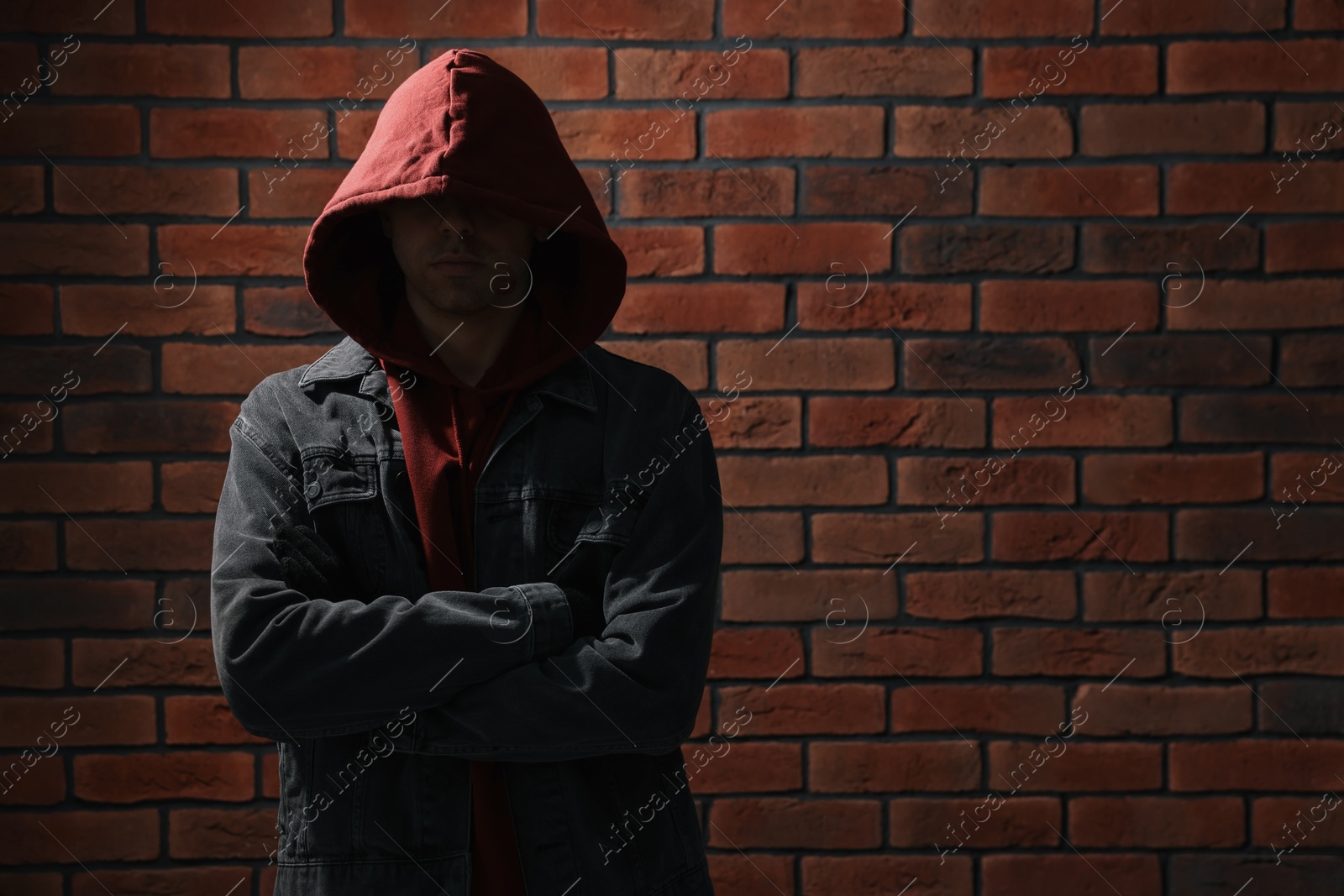 Photo of Thief in hoodie with crossed arms against red brick wall. Space for text