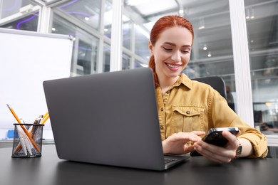 Photo of Happy woman with smartphone working on laptop at black desk in office
