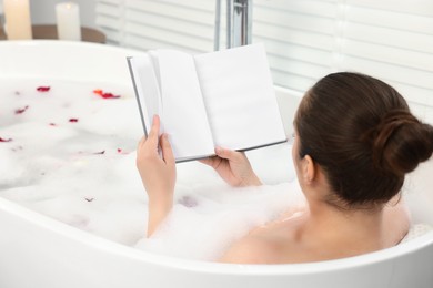 Photo of Woman reading book while taking bath in tub with foam and rose petals indoors, back view