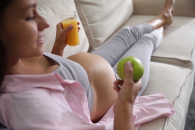 Photo of Young pregnant woman with apple and glass of juice in living room, closeup. Taking care of baby health