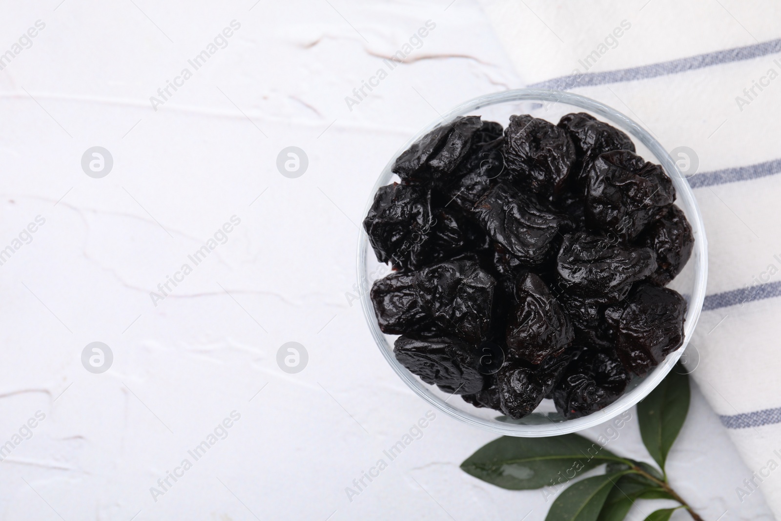 Photo of Sweet dried prunes in bowl and green leaves on white textured table, top view. Space for text