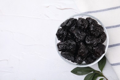 Sweet dried prunes in bowl and green leaves on white textured table, top view. Space for text
