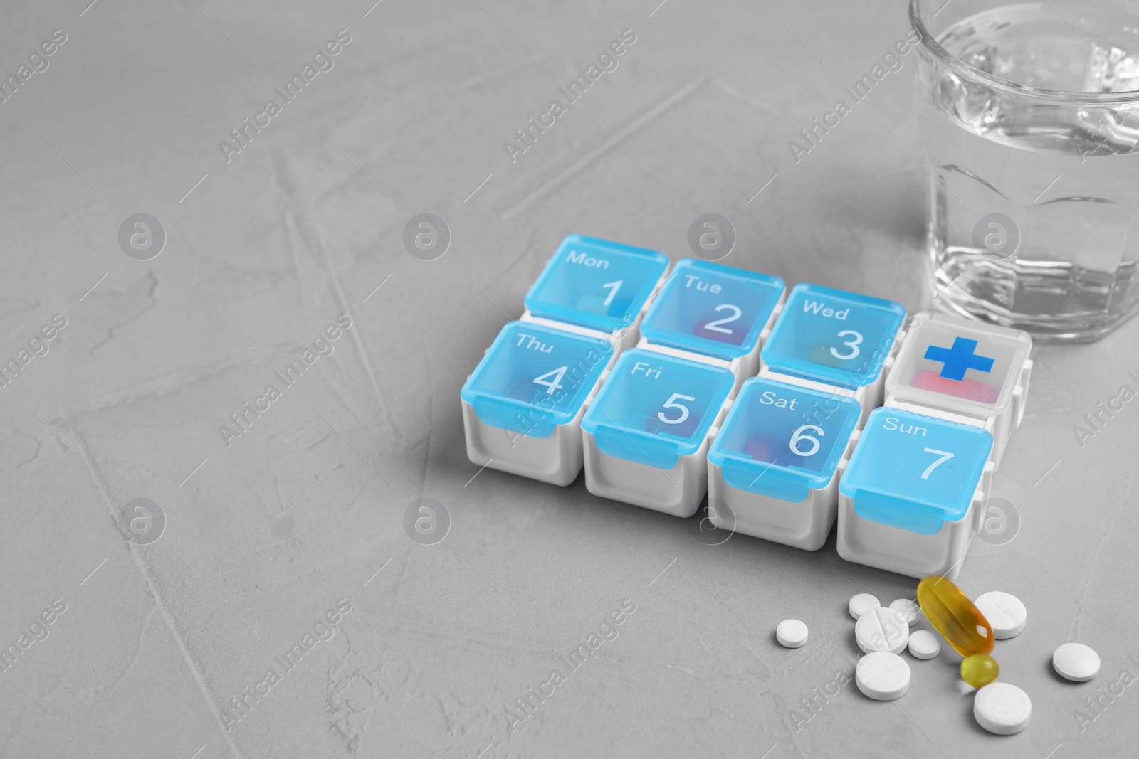 Photo of Weekly pill box with medicaments and glass of water on grey table. Space for text