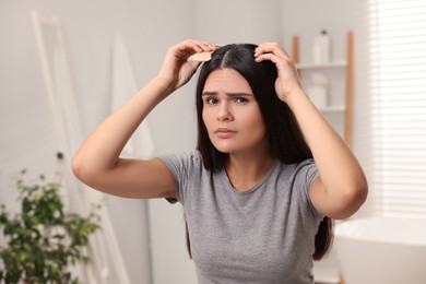 Emotional woman with comb examining her hair and scalp in bathroom. Dandruff problem