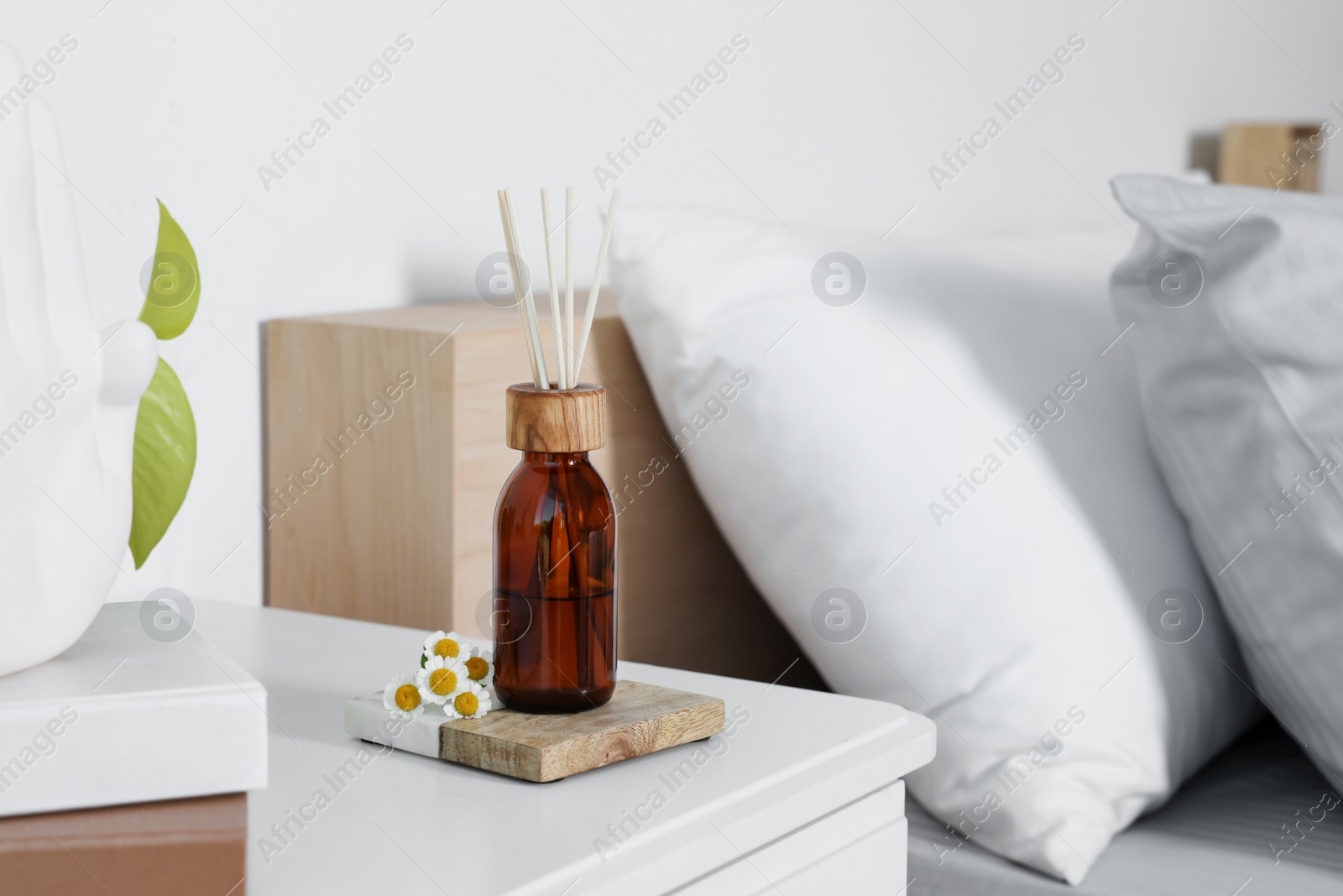 Photo of Aromatic reed air freshener and chamomiles on white nightstand in bedroom