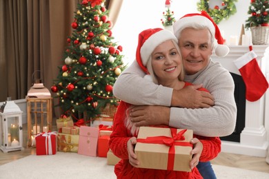 Happy mature couple in Santa hats with gift box at home, space for text. Christmas celebration
