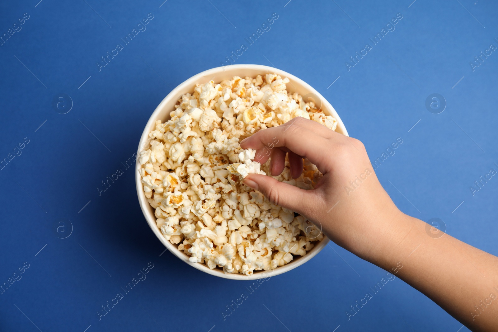 Photo of Woman taking fresh pop corn from bucket on blue background, top view