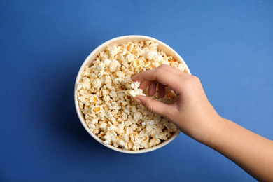 Woman taking fresh pop corn from bucket on blue background, top view