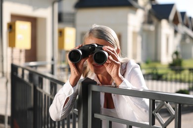 Photo of Concept of private life. Curious senior woman with binoculars spying on neighbours over fence outdoors