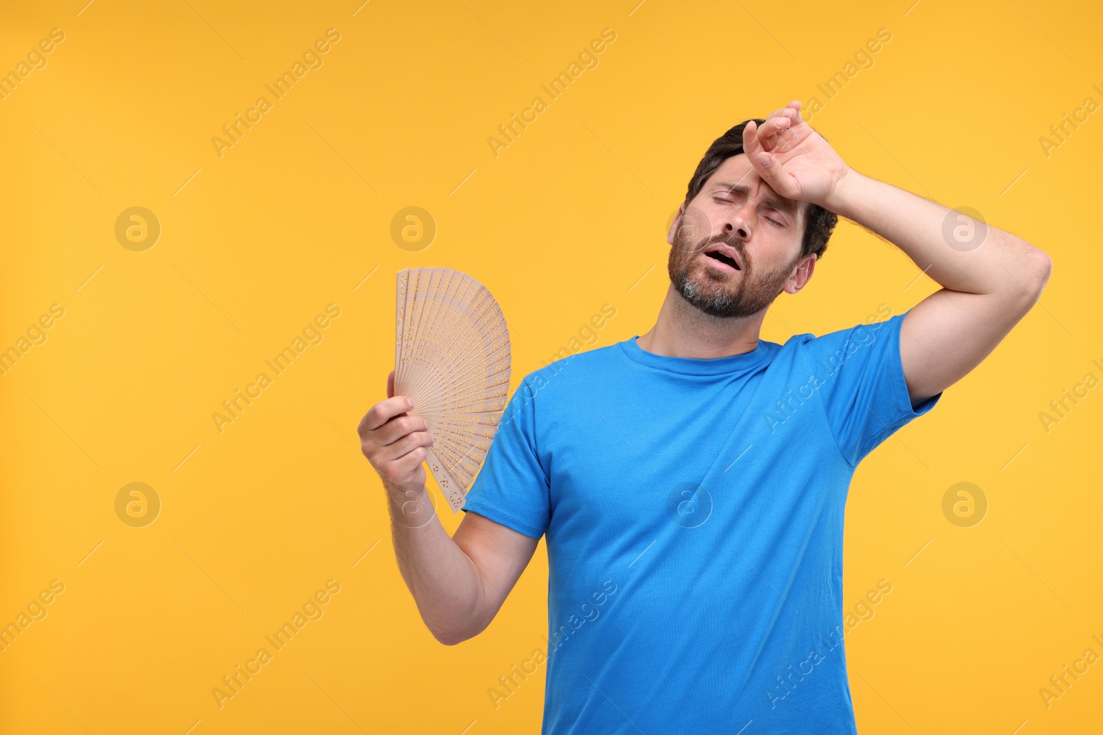 Photo of Unhappy man with hand fan suffering from heat on orange background. Space for text