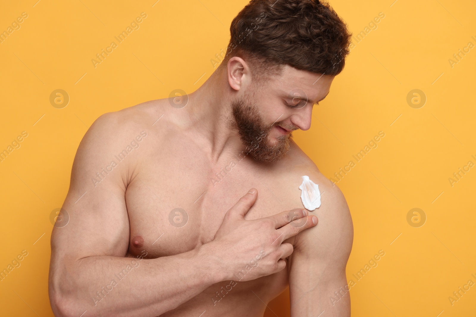 Photo of Handsome man applying moisturizing cream onto his shoulder on orange background