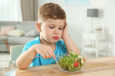 Photo of Sad little boy eating vegetable salad at table in room