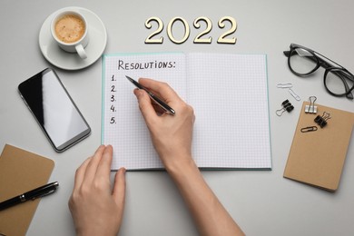 Photo of Woman filling list of resolutions for 2022 new year in notebook on light background, top view