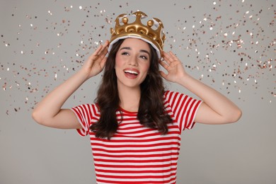 Beautiful young woman with inflatable crown under falling confetti on grey background
