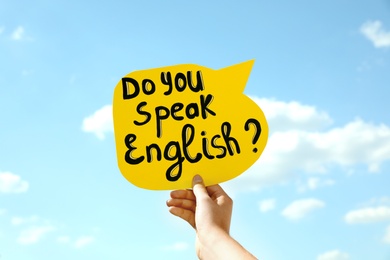 Photo of Woman holding paper speech bubble with question Do You Speak English against blue sky, closeup