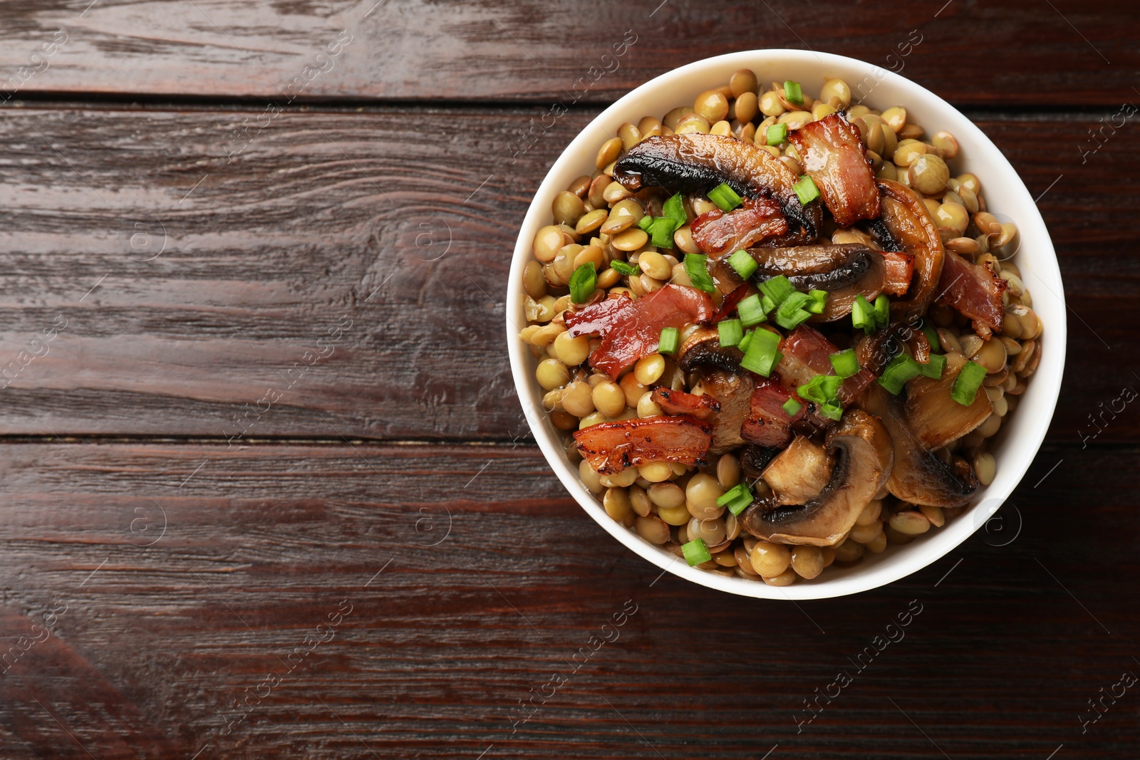 Photo of Delicious lentils with mushrooms, bacon and green onion in bowl on wooden table, top view. Space for text