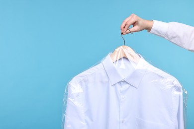 Photo of Dry-cleaning service. Woman holding shirt in plastic bag on light blue background, closeup. Space for text
