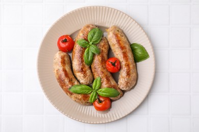 Photo of Plate with tasty homemade sausages, basil leaves and tomatoes on white tiled table, top view