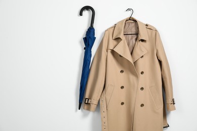 Photo of Hanger with beige trench coat and blue umbrella on white wall, space for text