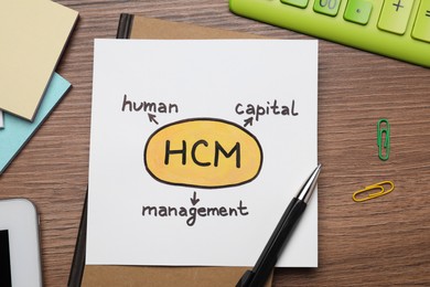 Paper with HCM abbreviation on wooden office table, flat lay. Human Capital Management