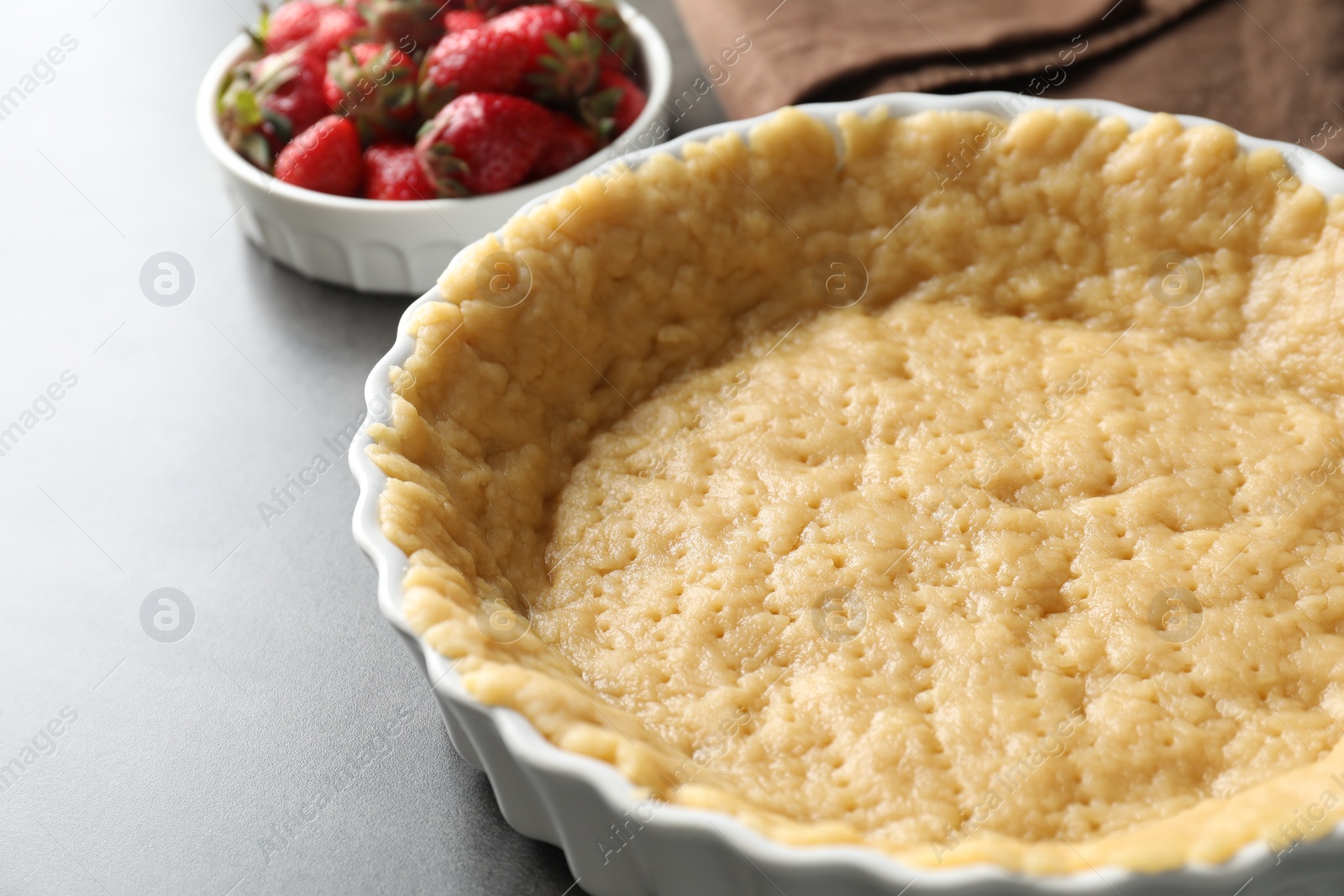 Photo of Making shortcrust pastry. Raw dough in baking dish and strawberries on grey table, closeup