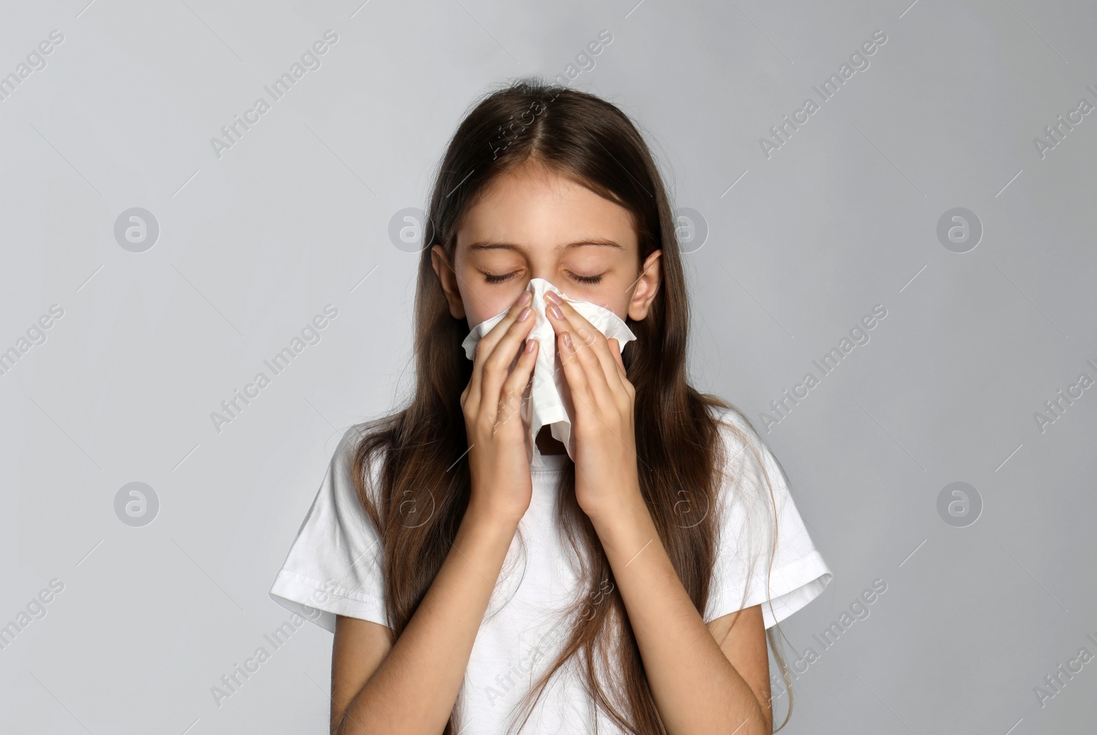 Photo of Little girl blowing nose into paper tissue on light grey background. Seasonal allergy