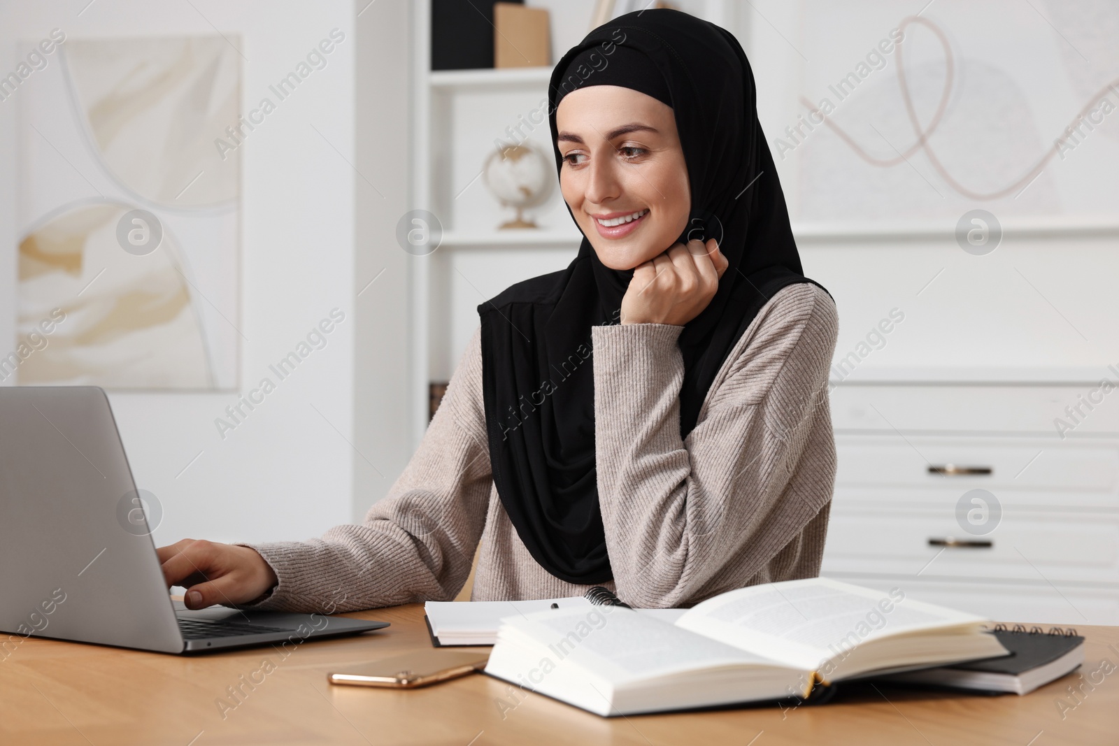 Photo of Muslim woman working near laptop at wooden table in room