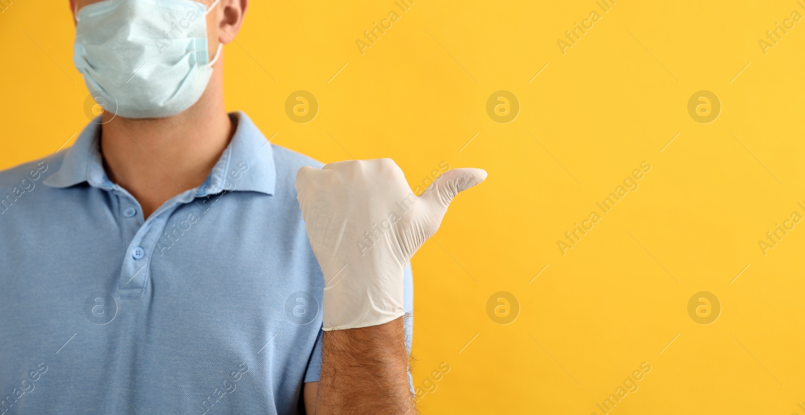 Photo of Man in protective face mask and medical gloves pointing at something on yellow background, closeup. Space for text