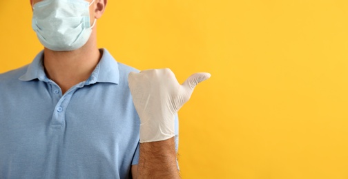 Man in protective face mask and medical gloves pointing at something on yellow background, closeup. Space for text