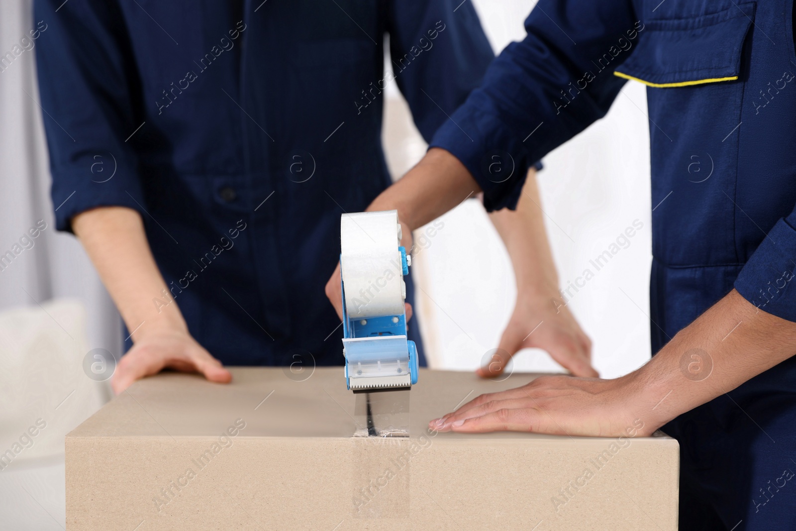 Photo of Male movers packing box with adhesive tape indoors, closeup