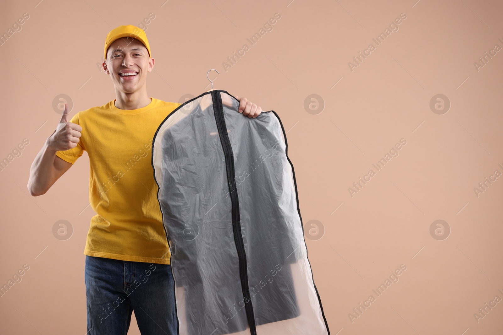 Photo of Dry-cleaning delivery. Happy courier holding garment cover with clothes and showing thumbs up on beige background, space for text