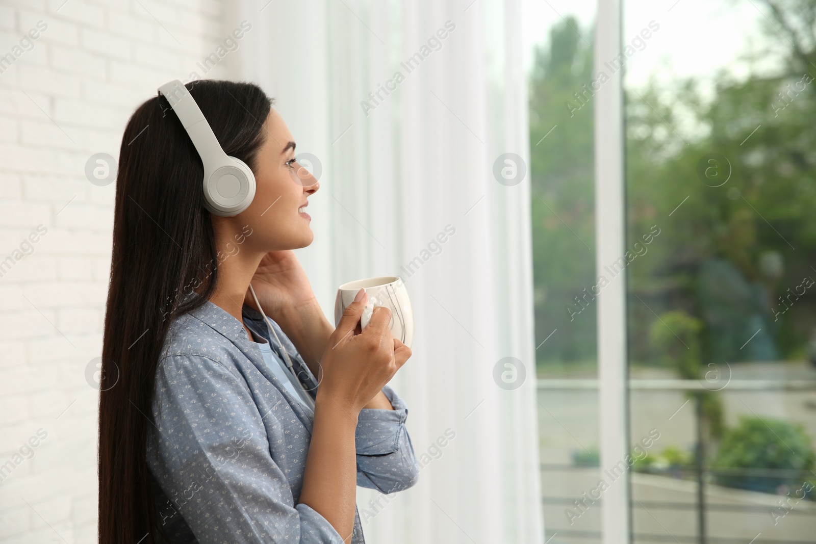 Photo of Young woman with cup of drink listening to music near window at home, space for text