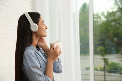 Young woman with cup of drink listening to music near window at home, space for text