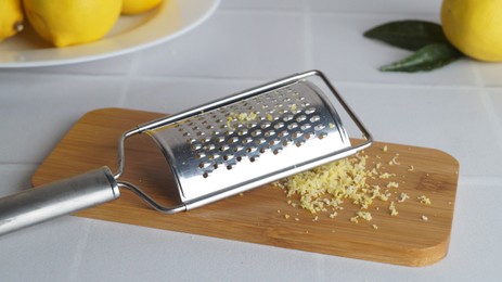 Wooden board with grater and fresh lemon zest on white tiled table