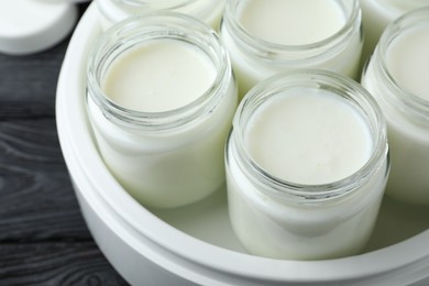 Photo of Modern yogurt maker with full jars on black wooden table, closeup