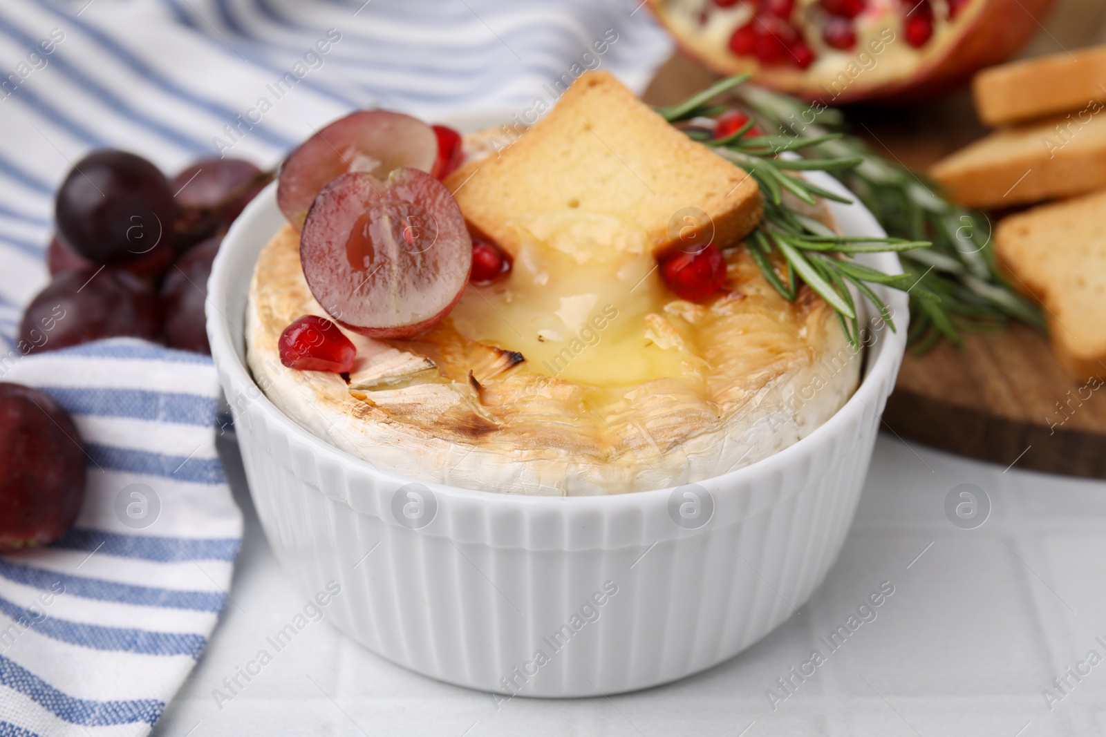 Photo of Tasty baked camembert with crouton, grape and rosemary on white tiled table, closeup