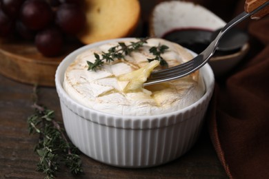 Photo of Eating tasty baked camembert with fork from bowl at wooden table, closeup