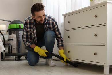 Photo of Man vacuuming floor under chest of drawers indoors