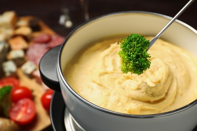 Photo of Dipping piece of broccoli into fondue pot with melted cheese on blurred background, closeup