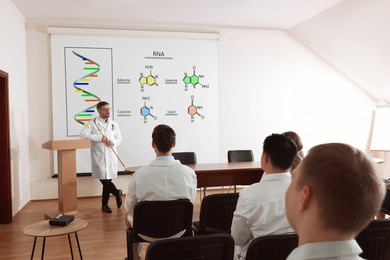 Image of Professor explaining audience nucleobases of RNA during lecture in conference room. Projection screen with illustration