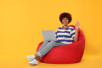 Photo of Beautiful young woman with laptop sitting on beanbag chair against orange background. Space for text