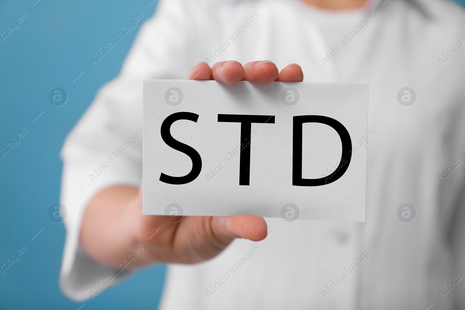 Photo of Doctor holding sheet of paper with abbreviation STD on light blue background, closeup